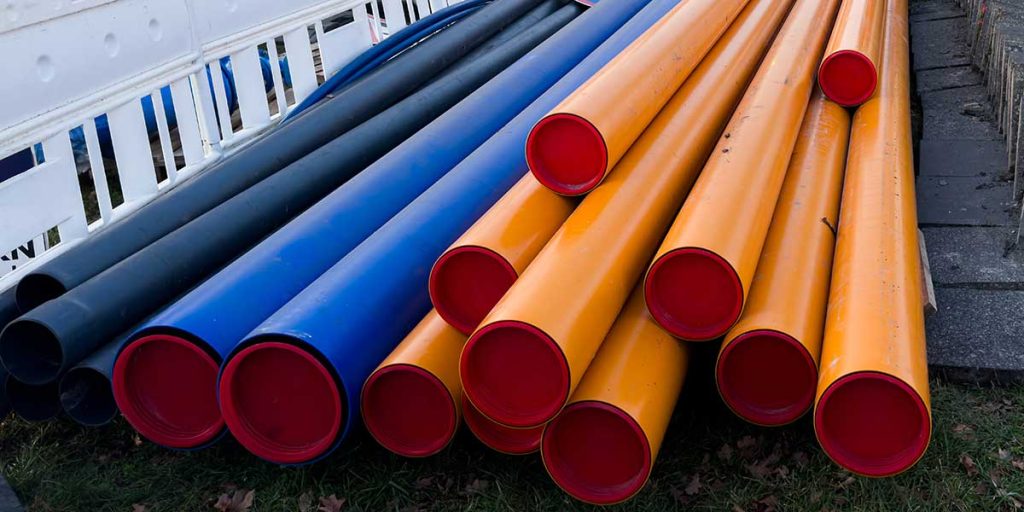 A large stack of different colored plumbing pipes including yellow, blue, and black options piled outdoors on the ground.
