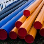 A large stack of different colored plumbing pipes including yellow, blue, and black options piled outdoors on the ground.