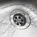 Water pools and swirls slowly around a stainless steel sink drain, illustrating a household plumbing issue with multiple slow drains.