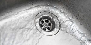 Water pools and swirls slowly around a stainless steel sink drain, illustrating a household plumbing issue with multiple slow drains.