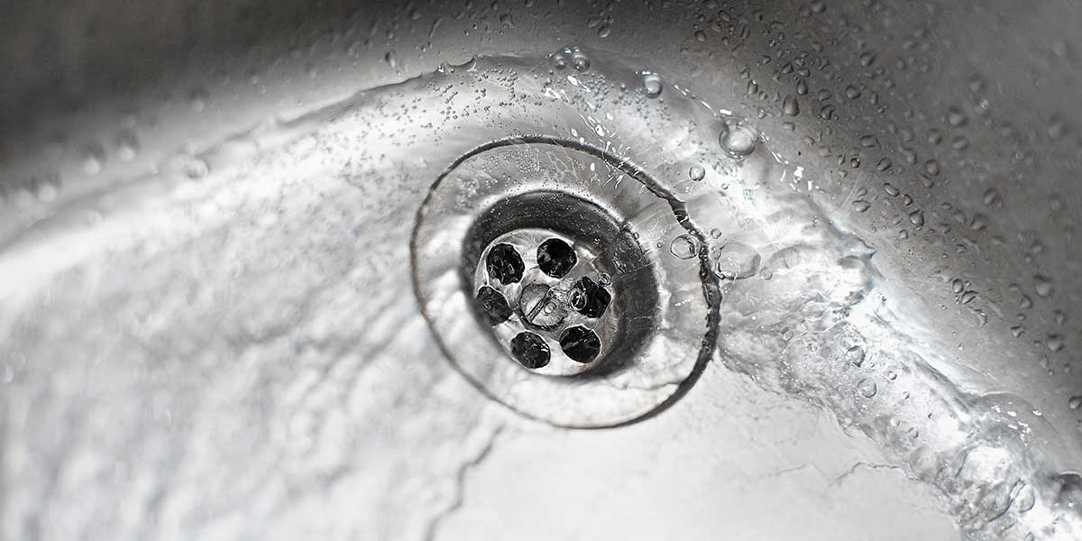Water pools and swirls slowly around a stainless steel sink drain, illustrating a household plumbing issue with multiple slow drains.