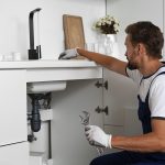 Professional plumber checking drainage pipes beneath a kitchen sink to diagnose plumbing problems.
