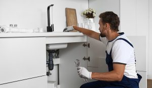 Professional plumber checking drainage pipes beneath a kitchen sink to diagnose plumbing problems.