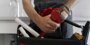 A plumber holds a large pipe wrench over an open tool bag containing pipes to prepare plumbing for heavy rain.
