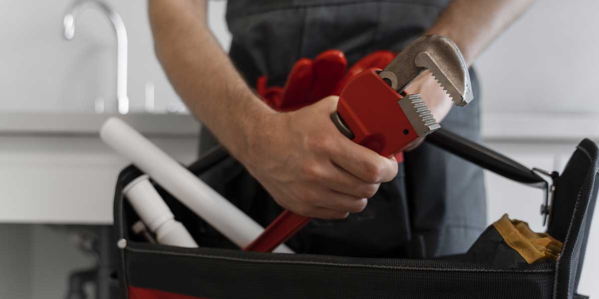 A plumber holds a large pipe wrench over an open tool bag containing pipes to prepare plumbing for heavy rain.